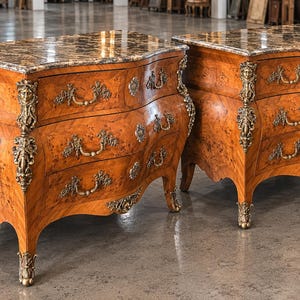 May include: A pair of wooden commodes featuring a brown burl wood finish, marble tops, and three drawers each. The furniture is adorned with ornate gold-toned metal hardware, including decorative drawer pulls and detailed corner accents.