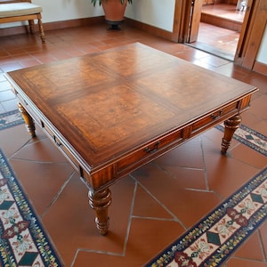 May include: A square, wooden coffee table with a rich, dark brown finish. The tabletop features a patterned design and is framed by a darker border. The table has four drawers and is supported by four ornate, turned legs. A classic piece of furniture.