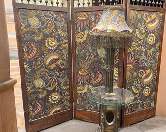 May include: A three-panel folding room divider with dark wood frames and fabric inserts featuring a colorful floral and pineapple pattern, paired with a tall table lamp that has a glass top and a fringed, patterned fabric lampshade.