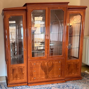 May include: A large, wooden display cabinet with glass doors and shelves. The cabinet has a warm, reddish-brown finish and features decorative details on the doors and drawers. The cabinet has three drawers and a heart-shaped design on the center drawer.