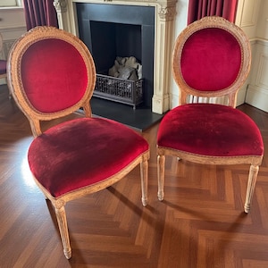 May include: Two antique chairs with oval backs and seats upholstered in deep red velvet. The wooden frames have a light finish with carved details. The chairs are set against a backdrop of a fireplace and hardwood flooring.