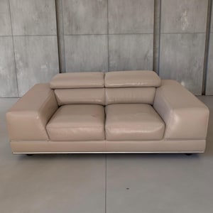 May include: A beige leather loveseat with a modern design. The two-seater sofa has a low profile, with a rectangular shape and a padded back and armrests. The sofa sits on a low, silver-colored base and has a smooth, clean appearance.
