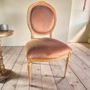 May include: An antique chair with a light wood frame and a plush, oval-shaped back and seat in a muted rose color. The chair has ornate carvings and delicate legs, showcasing a classic design.