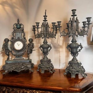 May include: An antique bronze clock and two candelabras are displayed on a wooden surface. The clock features a round face and cherub figures. The candelabras have multiple arms for candles.