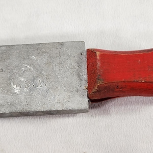 May include: A metal lead mold with a red wooden handle. The mold has a small metal pin and the text "U.S.A." is visible on the mold.