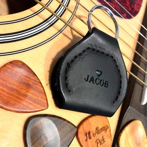 Puede incluir: Un porta-púas de guitarra de cuero negro con un llavero plateado y el nombre "JACOB" grabado en él. También se muestran cuatro púas de guitarra de madera, una con el texto "I'll Always Pick U!"