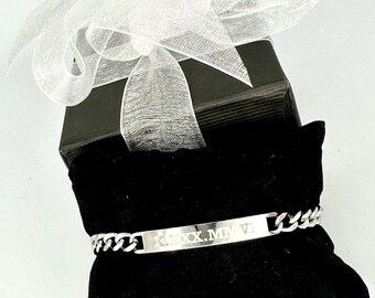 May include: A silver chain bracelet with a rectangular tag engraved with the Roman numeral date "XXXX.MMVI". The bracelet is displayed on a black velvet cushion inside a black gift box with a white ribbon.