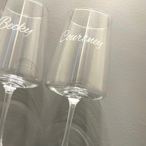 May include: Two clear champagne flutes with slender stems. The name "Becky" is etched on one glass, and "Courtney" on the other. The glasses are angled slightly, showcasing their elegant design.