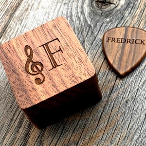 May include: A wooden guitar pick and a wooden box with a treble clef and the letter "F" engraved on the top. The guitar pick has the name "FREDRICK" engraved on it. The box is dark brown and the pick is a lighter brown.