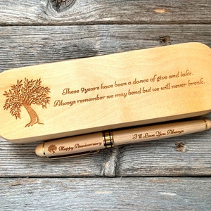 Puede incluir: Un juego de pluma y estuche de madera. El estuche está grabado con un diseño de árbol y el texto "These 9 years have been a dance of give and take." La pluma está grabada con "Happy Anniversary" y "I'll Love You Always."