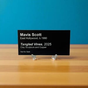 May include: A black sign with white text, supported by two clear plastic stands, against a blue backdrop. The sign displays "Mavis Scott, East Hollywood, b.1990, Tangled Vines, 2025, Clay Sculpture and Copper, Not for Sale."