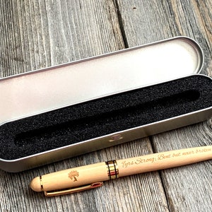 May include: A light-colored wooden pen with gold-colored accents and a matching case. The pen has the words "Tyrs Strong; Bent but never broken" engraved on the side. The pen case is silver and open, revealing a black foam interior.