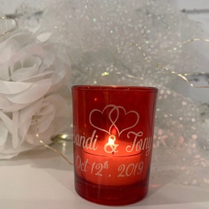 May include: Red glass votive candle holder with a white inscription that reads "Sandi & Tony Oct 12th, 2019" and two intertwined hearts.