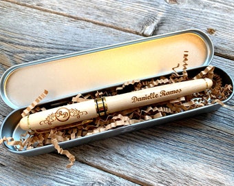 Puede incluir: Un bolígrafo de madera personalizado con detalles dorados, grabado con el nombre "Danielle Ramos" y las iniciales "RD" dentro de una corona decorativa. El bolígrafo está en un estuche metálico abierto lleno de virutas de papel marrón.