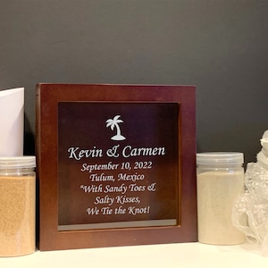 May include: A wooden framed sign with the names "Kevin & Carmen", date, and location. Two clear jars with sand and a white paper cone are on the left. A white floral arrangement is on the right.