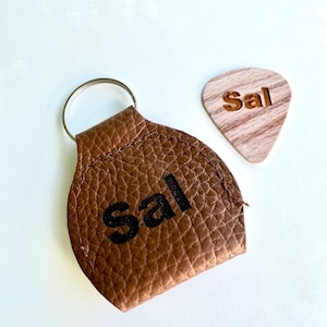 Puede incluir: Un llavero de cuero marrón con el nombre "Sal" impreso en negro. Al lado, una púa de guitarra de madera con el nombre "Sal" grabado. El llavero tiene un llavero plateado.