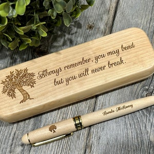 Può includere: Un set di penna e astuccio in legno chiaro. L'astuccio porta la citazione "Always remember, you may bend but you will never break" e un disegno di albero. La penna ha un disegno di albero e il nome "Havala McKinney".