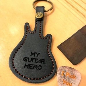 Puede incluir: Llavero de cuero negro con forma de guitarra con el texto "MY GUITAR HERO" y una púa que dice "I Pick You". El llavero tiene un anillo de color bronce y un emblema decorativo. La púa es de color marrón claro con texto naranja.