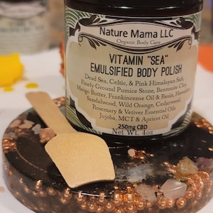 May include: A glass jar with a black label that reads "Nature Mama LLC Organic Body Care Vitamin "SEA" Emulsified Body Polish" with a list of ingredients including Dead Sea, Celtic, & Pink Himalayan Salt, Finely Ground Pumice Stone, Bentonite Clay, Mango Butter, Frankincense Oil & Resin, Hawaiian Sandalwood, Wild Orange, Cedarwood, Rosemary & Vetiver Essential Oils, Jojoba, MCT & Apricot Oil. The label also states "250mg CBD Net Wt. 4oz".
