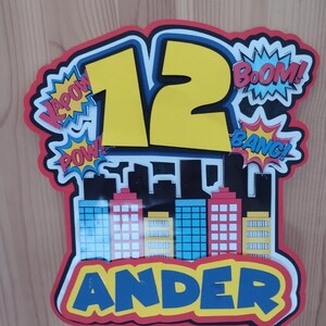 Puede incluir: Un adorno para tarta de cumpleaños colorido con temática de superhéroes. El número "12" es amarillo, rodeado de efectos de sonido de cómic como "Pow!" y "Boom!". Un paisaje urbano en la parte inferior, con el nombre "Ander" debajo.