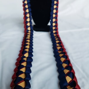 May include: A blue and burgundy lei made of ribbon, with a gold accent. The lei is braided and has a traditional Hawaiian design.