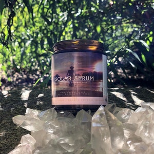 K&ouml;nnte beinhalten: Ein braunes Glas Solar Serum mit schwarzem Deckel, vor dem Hintergrund gr&uuml;ner Bl&auml;tter und klarer Quarzkristalle. Das Etikett zeigt eine Sonnenuntergangsszene und den Text "Nourishing Summer Skin". Das Glas enth&auml;lt ein Hautpflegeprodukt.