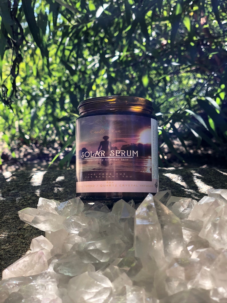 K&ouml;nnte beinhalten: Ein braunes Glas Solar Serum mit schwarzem Deckel, vor dem Hintergrund gr&uuml;ner Bl&auml;tter und klarer Quarzkristalle. Das Etikett zeigt eine Sonnenuntergangsszene und den Text "Nourishing Summer Skin". Das Glas enth&auml;lt ein Hautpflegeprodukt.