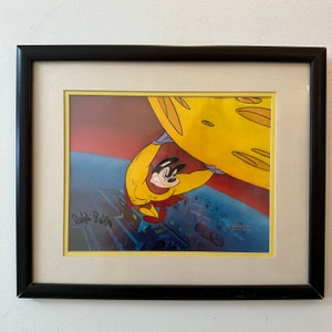 May include: A framed cartoon cel featuring a yellow and red superhero character holding up a large yellow disc. The character has a black nose and black eyes. The cel is signed "Ralph Bakshi" in the bottom left corner and "Remarkable" in the bottom right corner.