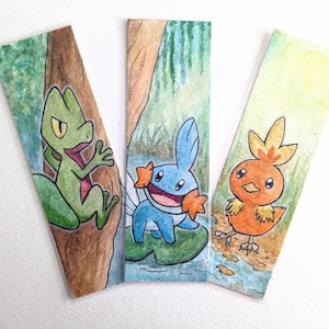 May include: Three watercolour bookmarks featuring Pokemon characters. The first bookmark shows a green lizard-like Pokemon with a yellow eye, the second bookmark shows a blue water-type Pokemon with a light blue fin, and the third bookmark shows an orange bird-like Pokemon with a yellow beak.