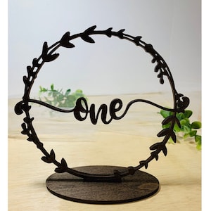 May include: A wooden table number with a black wreath design and the word "one" in a cursive font. The table number is on a wooden base.