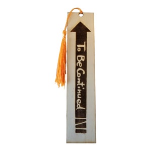 May include: Wooden bookmark with a brown arrow pointing upwards and the text "To Be Continued" in black lettering. The bookmark has a tassel made of orange string.
