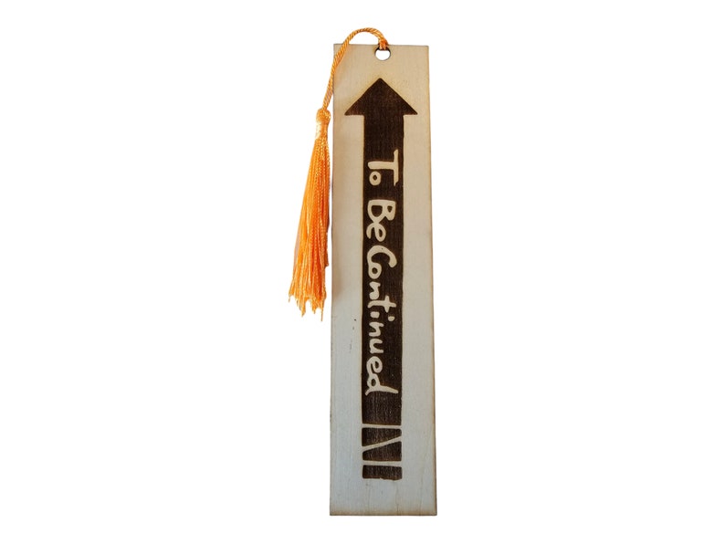 May include: Wooden bookmark with a brown arrow pointing upwards and the text "To Be Continued" in black lettering. The bookmark has a tassel made of orange string.