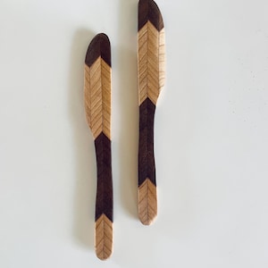 May include: Two wooden butter knives with a chevron pattern. The handles and tips are dark brown, while the center features a light wood chevron design. The knives are displayed on a white surface.
