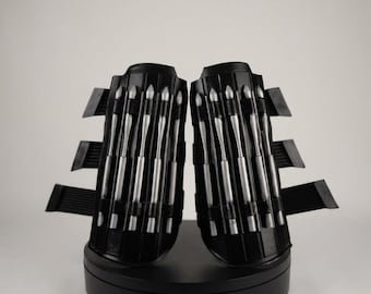 May include: A pair of black forearm guards featuring three vertical silver-toned metallic spikes on each piece. The guards have a curved, rigid structure with three horizontal black straps extending from the outer edges of each side.