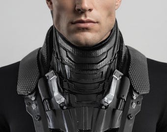 May include: A black neck and shoulder armor piece featuring a segmented, high-collar design with textured panels and metallic silver accents. The structure is rigid, covering the neck and extending over the shoulders, worn over a plain black shirt.