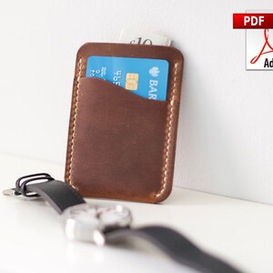 May include: A brown leather cardholder with a light-colored stitched border. A blue credit card and a folded bill are visible inside. A black wristwatch with a silver face and black band is in the foreground.