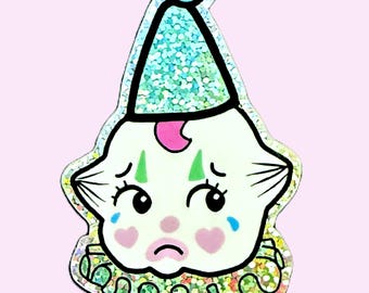 May include: A white, cartoon-style clown face with green eyebrows, pink cheeks, and blue teardrop-shaped eyes. The clown is wearing a blue and green glitter party hat and a green and white glitter collar.