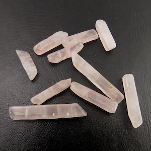 May include: A collection of pale pink rose quartz crystal wands. The wands vary in size and shape, with a translucent appearance. The crystals are scattered on a dark surface, showcasing their natural beauty and subtle color variations.