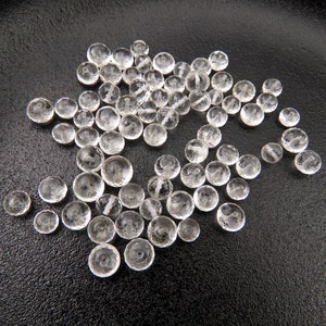 K&ouml;nnte beinhalten: Eine Sammlung von klaren, facettierten Glasperlen, die auf einer schwarzen Oberfl&auml;che verstreut sind. Die Perlen sind rund und haben eine leicht unregelm&auml;&szlig;ige Form.