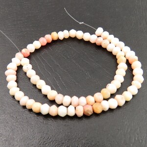 May include: Strand of faceted, light pink opal beads. The beads are strung on a thin, clear wire, and the color varies from white to peach. The beads are small and have a geometric shape. The background is a dark gray.