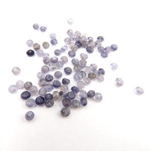 May include: A collection of small, faceted beads in varying shades of blue and grey. The beads are round and appear translucent, with a polished finish. They are scattered on a white background, creating a delicate display.
