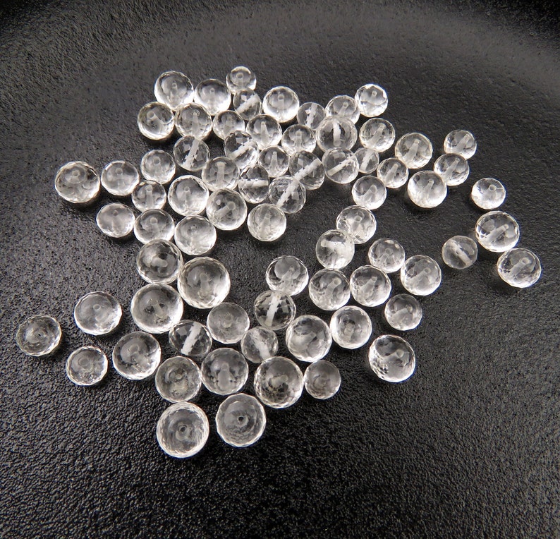 K&ouml;nnte beinhalten: Eine Sammlung von klaren, facettierten Glasperlen, die auf einer schwarzen Oberfl&auml;che verstreut sind. Die Perlen sind rund und haben eine leicht unregelm&auml;&szlig;ige Form.