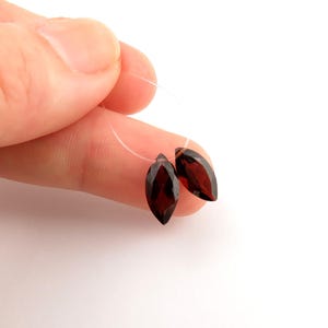 May include: Two faceted, dark red marquise-shaped gemstones strung on a thin, clear thread. The gemstones are held against a white background, showcasing their deep color and cut. Ideal for jewelry making.