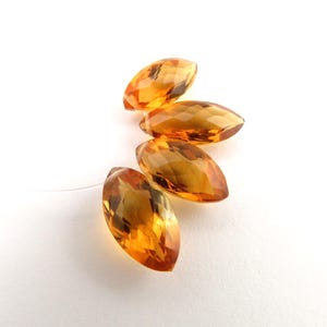 May include: Five faceted, marquise-cut gemstones in shades of amber and gold are displayed against a white background. The gems are strung together on a thin, clear thread, suggesting they are part of a jewelry design or craft project.