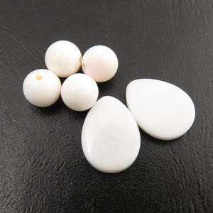 May include: A collection of white beads and egg-shaped cabochons. The beads are round, with one having a hole. The cabochons are smooth and oval, with a slight curve. The objects are arranged on a dark, textured surface.