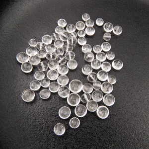 K&ouml;nnte beinhalten: Eine Sammlung von klaren, facettierten Glasperlen, die auf einer schwarzen Oberfl&auml;che verstreut sind. Die Perlen sind rund und haben eine leicht abgeflachte Form.