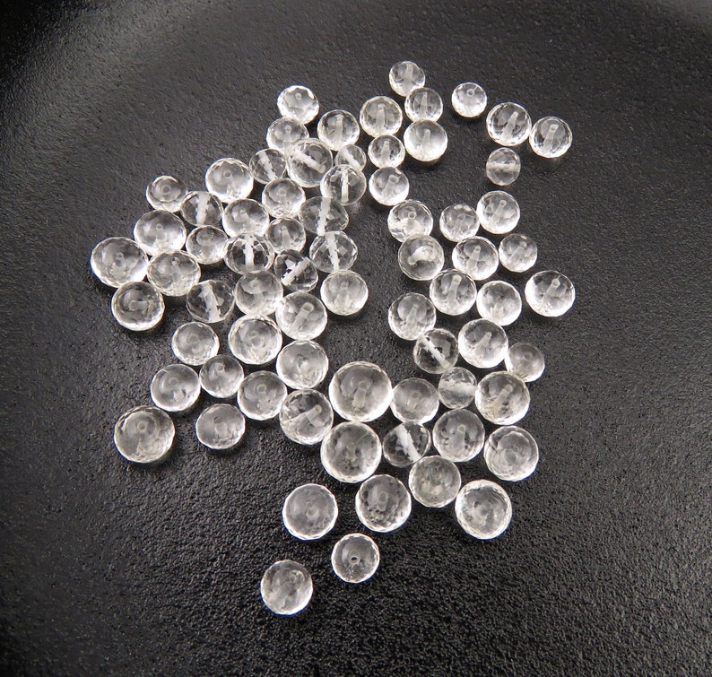 K&ouml;nnte beinhalten: Eine Sammlung von klaren, facettierten Glasperlen, die auf einer schwarzen Oberfl&auml;che verstreut sind. Die Perlen sind rund und haben eine leicht abgeflachte Form.