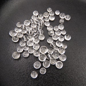 K&ouml;nnte beinhalten: Eine Sammlung von klaren, facettierten Glasperlen, die auf einer schwarzen Oberfl&auml;che verstreut sind. Die Perlen sind rund und haben eine leicht abgeflachte Form.