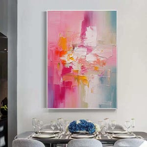 K&ouml;nnte beinhalten: Abstraktes &Ouml;lgem&auml;lde mit rosa, orange und wei&szlig;en Farbt&ouml;nen. Das Kunstwerk zeigt strukturierte Pinselstriche, die eine dynamische Komposition bilden. Das Gem&auml;lde ist wei&szlig; gerahmt und h&auml;ngt &uuml;ber einem Esstisch.