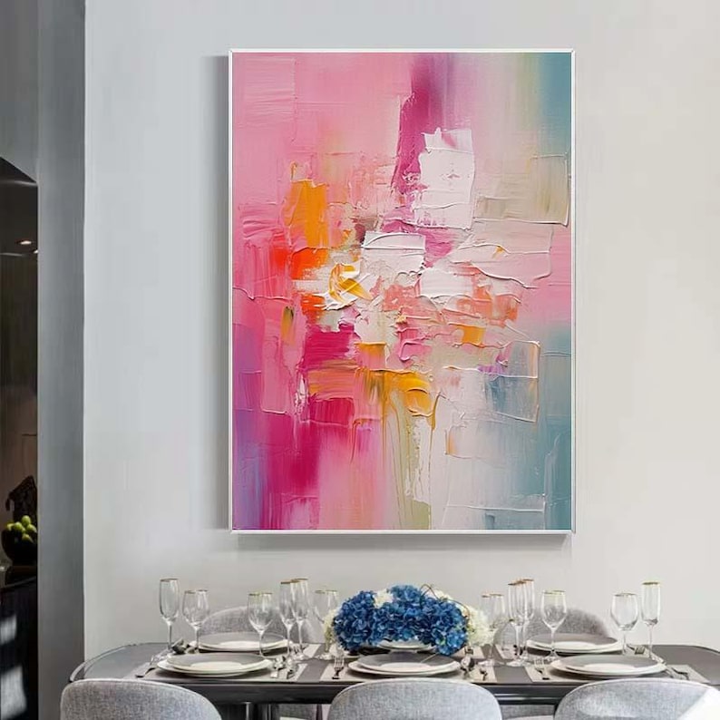 K&ouml;nnte beinhalten: Abstraktes &Ouml;lgem&auml;lde mit rosa, orange und wei&szlig;en Farbt&ouml;nen. Das Kunstwerk zeigt strukturierte Pinselstriche, die eine dynamische Komposition bilden. Das Gem&auml;lde ist wei&szlig; gerahmt und h&auml;ngt &uuml;ber einem Esstisch.